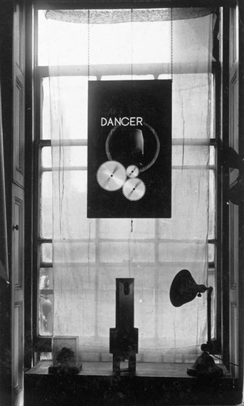 A black-and-white interior scene featuring a hanging poster with mechanical imagery and abstract objects on a windowsill.