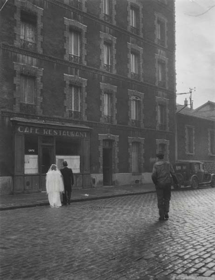 A black-and-white street scene captures a couple walking past a café while a soldier walks away in the foreground.