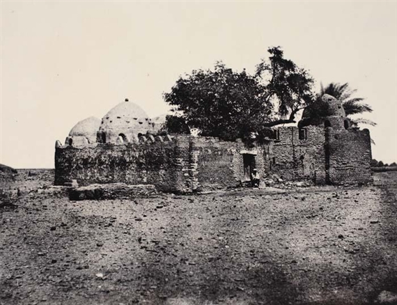 A sepia-toned photograph of a weathered stone structure with domes and crenellated walls, partially obscured by trees.
