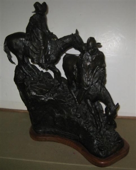 A dark bronze sculpture of two figures on horseback mounted on a wooden base.