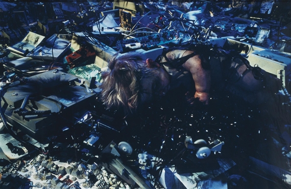 A figure lies amidst a chaotic pile of discarded electronics and debris.
