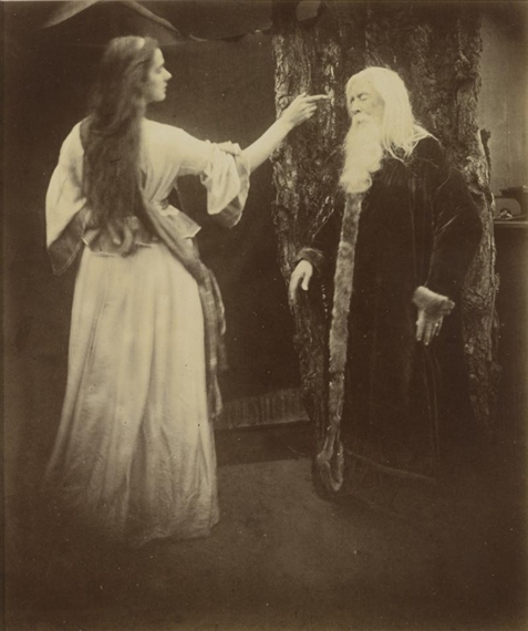 A sepia-toned theatrical scene featuring a woman in a flowing dress offering something to an elderly figure with a long white beard.
