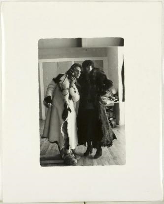 A black-and-white photograph capturing two figures in heavy winter coats standing close together indoors.