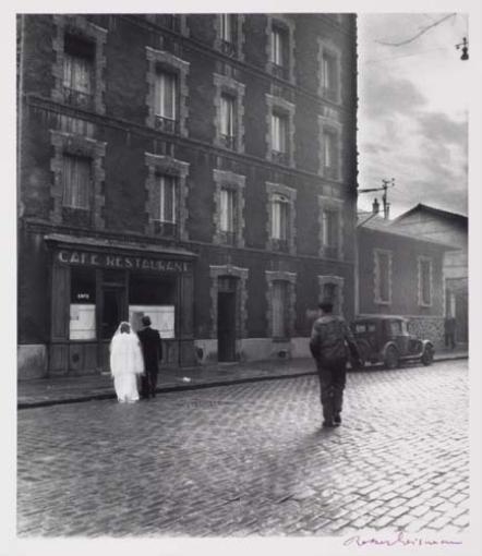 A black-and-white street scene captures a couple in formal wear standing before a café, with a solitary figure walking away down the cobblestone road.