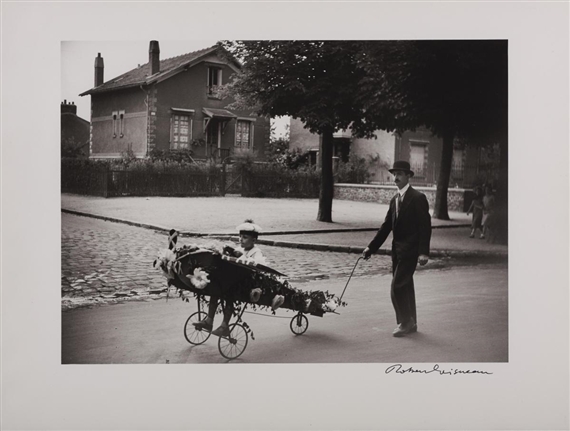 A black-and-white photograph captures a man pulling a flower-decorated cart with a seated figure inside, set against a quiet residential street.