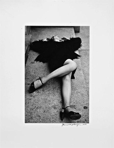 A black-and-white photograph of a person lying on a stone surface, wearing a dark feathered garment and high-heeled shoes.