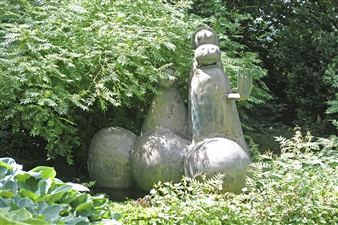 A group of abstract, rounded figures made of weathered metal stand amidst dense green foliage.