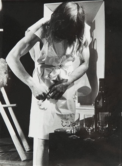 A black-and-white portrait of a person in a dress, bent over a table with bottles and glasses.