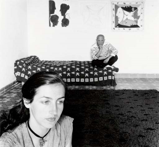 A black and white portrait featuring a woman in the foreground and a man sitting on a bed in the background.