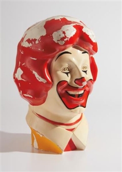 A weathered clown head bust with a vibrant red wig and peeling paint against a plain background.