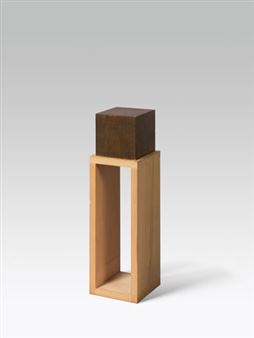 A minimalist sculpture featuring a light wooden frame supporting a dark cube.