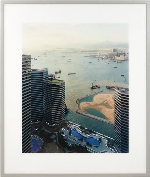 A high-angle view of a modern waterfront city with skyscrapers, pools, and a harbor filled with boats.
