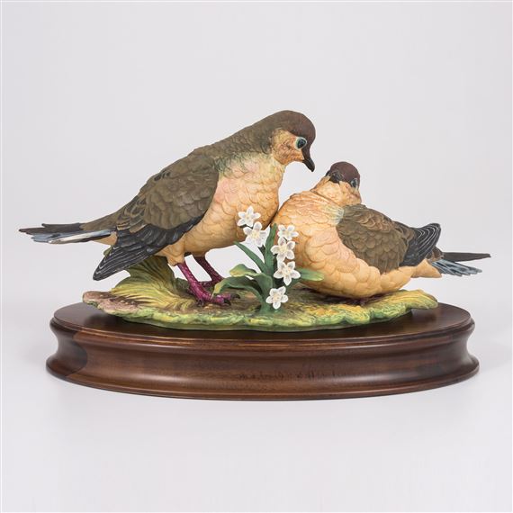 A sculptural diorama featuring two birds perched on a floral base atop a polished wooden plinth.
