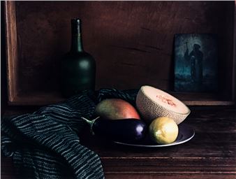 A moody still life featuring dark green glass, purple eggplant, and a halved melon on a plate.