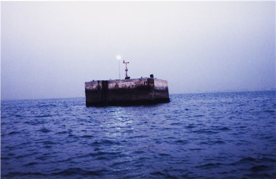 A solitary concrete block floats on a vast, rippling sea under a pale sky.