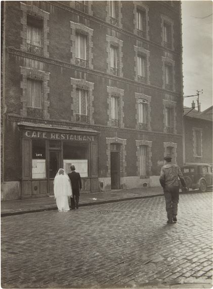 A vintage street scene captures a wedding couple entering a café while a soldier walks away on a cobblestone road.