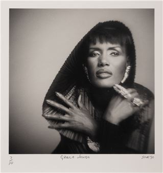 A black-and-white portrait of a person wearing a textured hooded garment, adorned with large jewelry and rings.