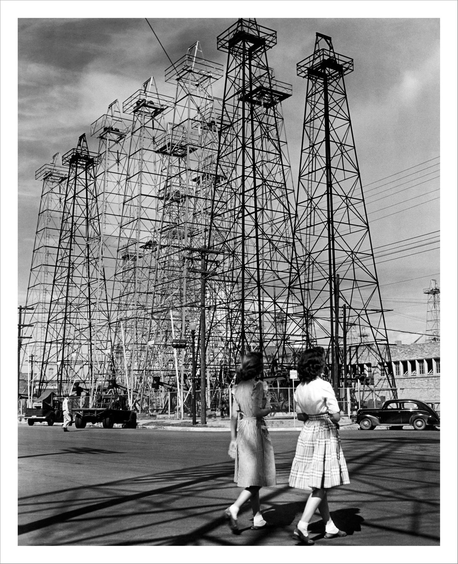 Andreas Feininger Two female walkers pass a row of large derricks in