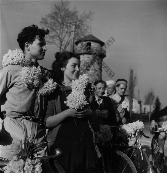 A black-and-white group portrait of a couple with flower garlands, standing before a windmill.