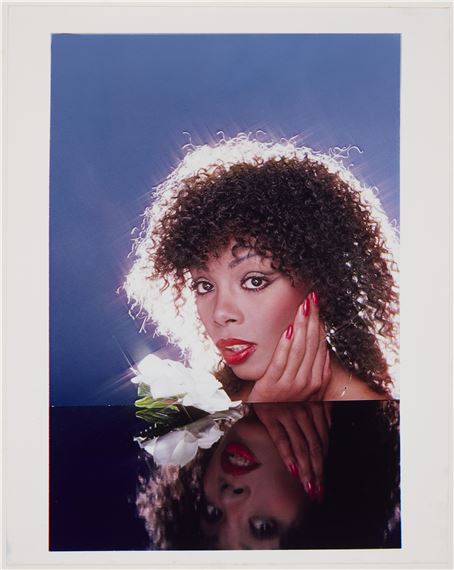 A portrait of a person with voluminous curly hair, red lipstick, and a white flower held near the face, reflected in a dark surface below.