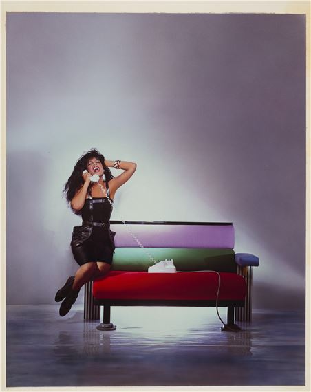 A person in a black outfit sits on a colorful sofa while holding a white telephone receiver.