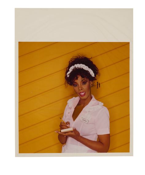 A portrait of a person in a pink uniform holding a notepad against a yellow slatted wall.