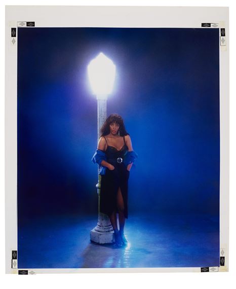 A portrait of a figure in a dark dress standing beside a glowing street lamp against a deep blue background.
