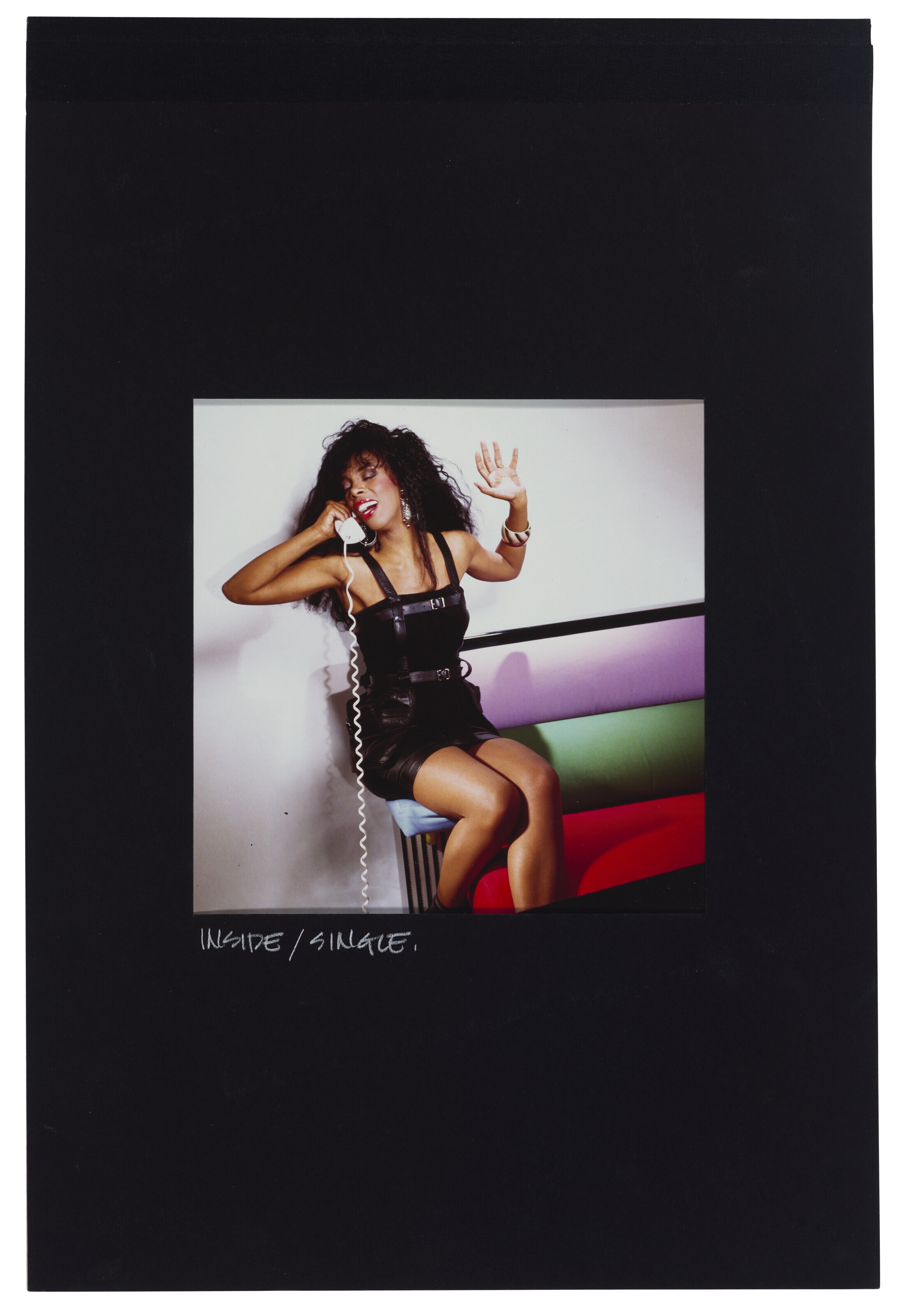 A candid portrait of a person in a black outfit sitting on a colorful bench while holding a telephone receiver.