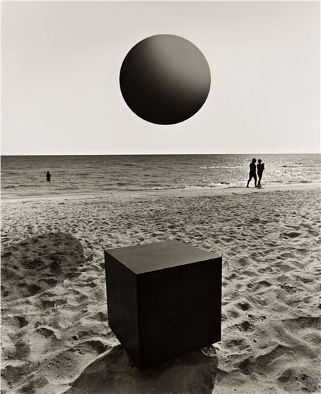 A surreal black-and-white beach scene featuring a large cube on the sand, an oversized sphere in the sky, and distant figures by the water.