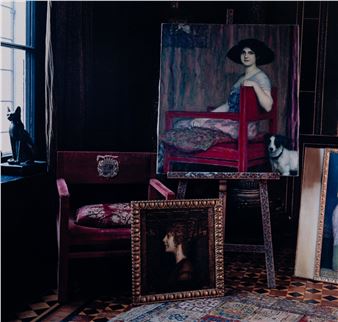 A moody interior scene featuring a red velvet chair, a large portrait on an easel, and smaller framed artworks.