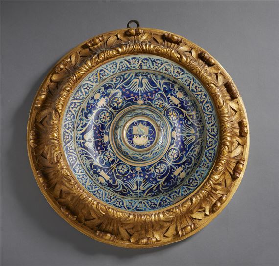 A painted and glazed maiolica plate, cavetto decorated with trophy and phytomorphic motifs on a blue background with umbo centered by a coat of arms, brim with a band alternating with stylized flowers - Master Giorgio