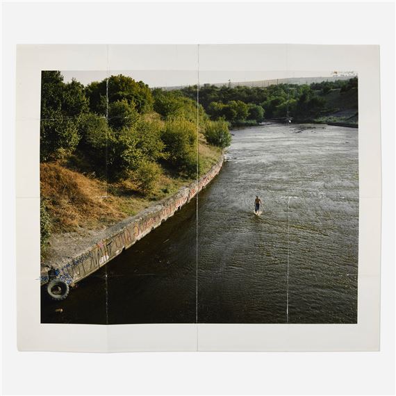 A high-angle view of a river with a solitary figure standing in the water, framed by a concrete embankment and distant hills.