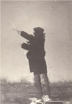 A vintage photograph captures a figure in profile, holding a long object high above their head against an open sky.