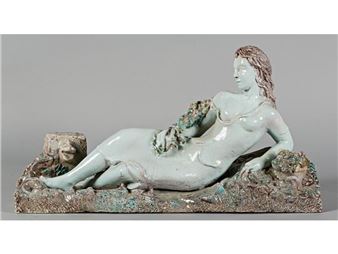 A pale green reclining figure rests on a textured, rocky base with a small face sculpture nearby.