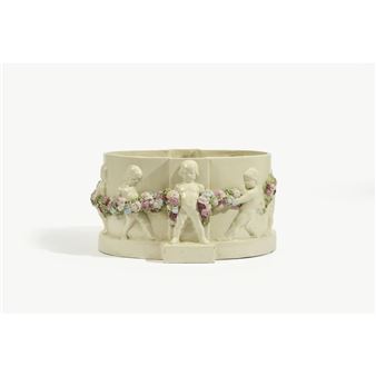 Jardinière-centre de table ovale reposant sur une haute base débordante, en faïence émaillée crème à décor en haut-relief de putti tenant une guirlande de fleurs émaillée polychrome. Signée “G. Dengg” au-dessus de la base. Cachet de la Manufacture Wiener Kunstkeramische Werkstätte au revers de la base “Conçu pour la société autrichienne pour la promotion de l’art des médailles et de la petite sculpture par l’Atelier de céramique artistique de Vienne”. - Gertrude Dengg