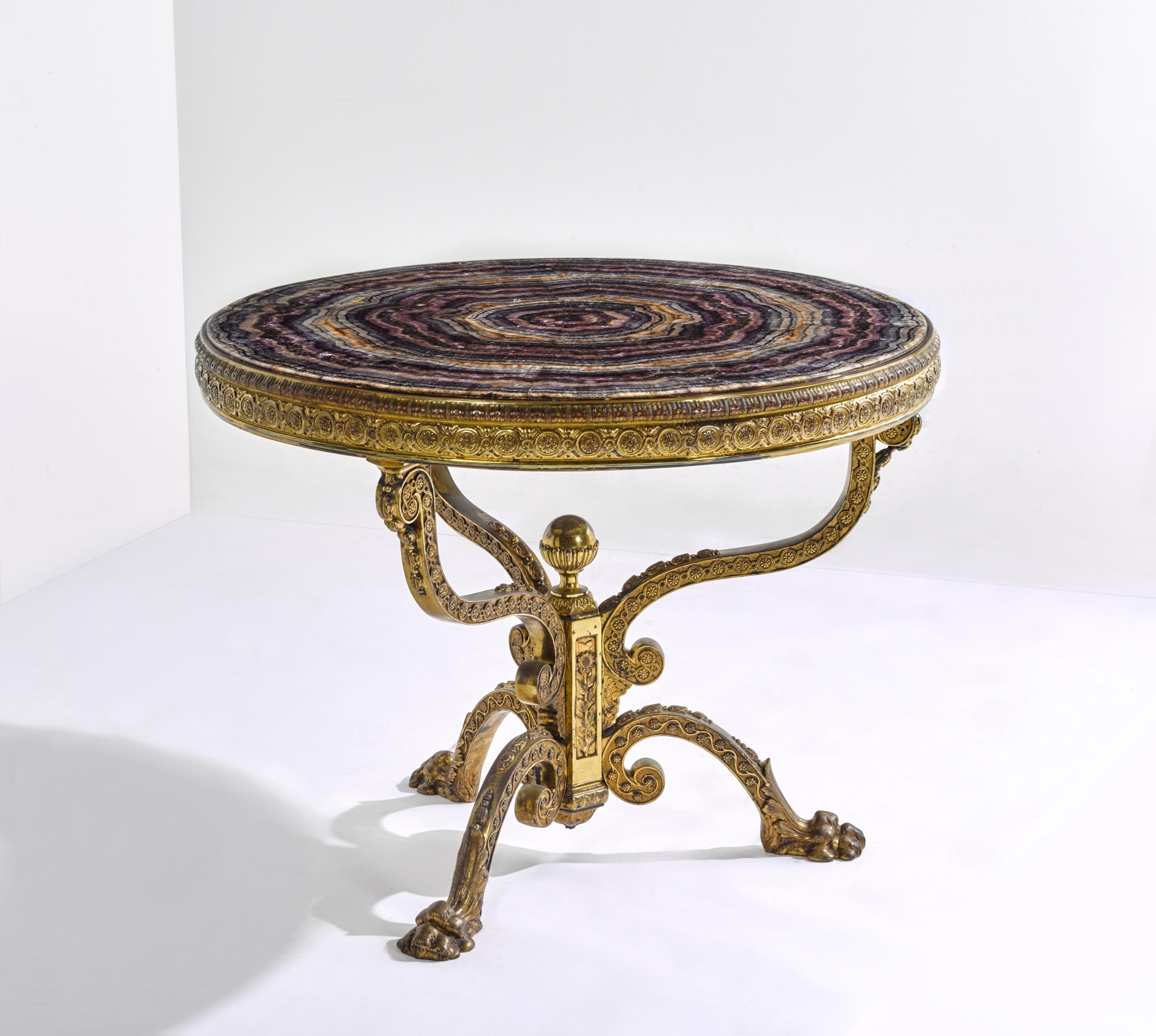 Artwork by Thomas Woodruff, A Victorian gilt-brass mounted Blue John centre table, , the top attributed to Thomas Woodruff, the base by William Tonks and Sons, Made of gilt-brass, the circular Blue John veneered top applied in segments to form concentric rings, the frieze with a border of paterae filled guilloche headed by fluting and stop-fluting, the three similarly cast scrolled supports decorated with husks and converging on a hexagonal pillar with a gadrooned ball finial and decorated on each facet with sunflowers, the arched tripod base similarly decorated with husks and trailing flowers on leafy paw feet