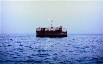 A solitary concrete block floats in a vast, calm sea under a pale sky.