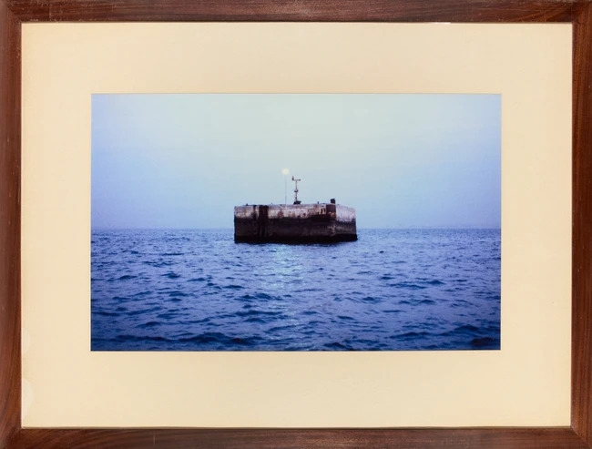 A solitary concrete block floats in a vast, calm sea under a pale sky.