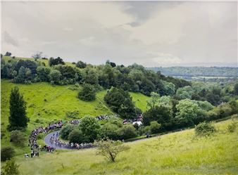 A sweeping landscape view of a winding road cutting through rolling green hills, where a large group of figures gathers along the curve.