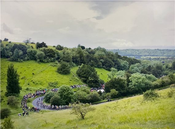 A sweeping landscape view of a winding road cutting through rolling green hills, where a large group of figures gathers along the curve.