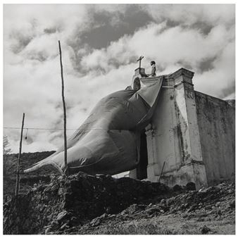 A black and white photograph of a person sitting on the roof of a church with a large, inflated object draped over its side.