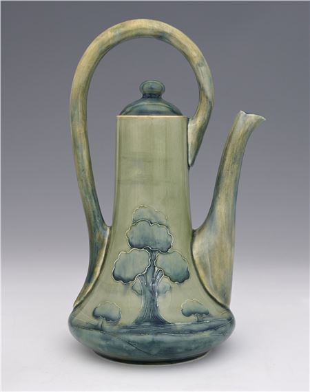 A tall ceramic pitcher with a greenish-blue glaze featuring a raised tree landscape design.