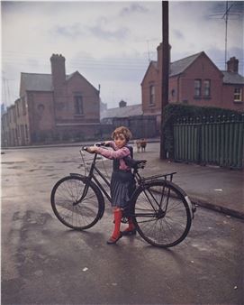 A vintage photograph of a young girl standing with a bicycle on a wet street.