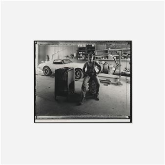 A black-and-white photograph of a person leaning against a tire in a garage, with a classic car in the background.