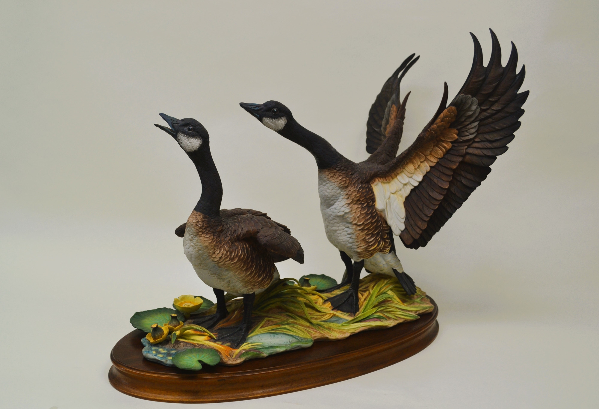 A sculptural diorama featuring two geese in flight and standing poses on a lily pad base.