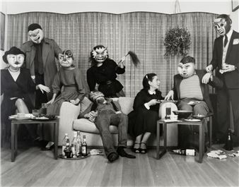 A surreal black-and-white group portrait featuring seven figures in a living room setting, all wearing bizarre masks.