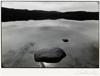 A black and white photograph of a calm lake with floating rocks reflecting the distant hills.