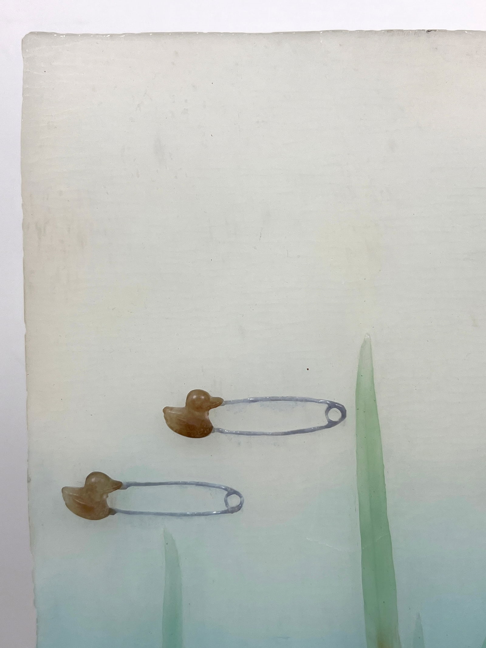 A vertical mixed-media artwork featuring tall green blades and scattered safety pins against a blue gradient background.