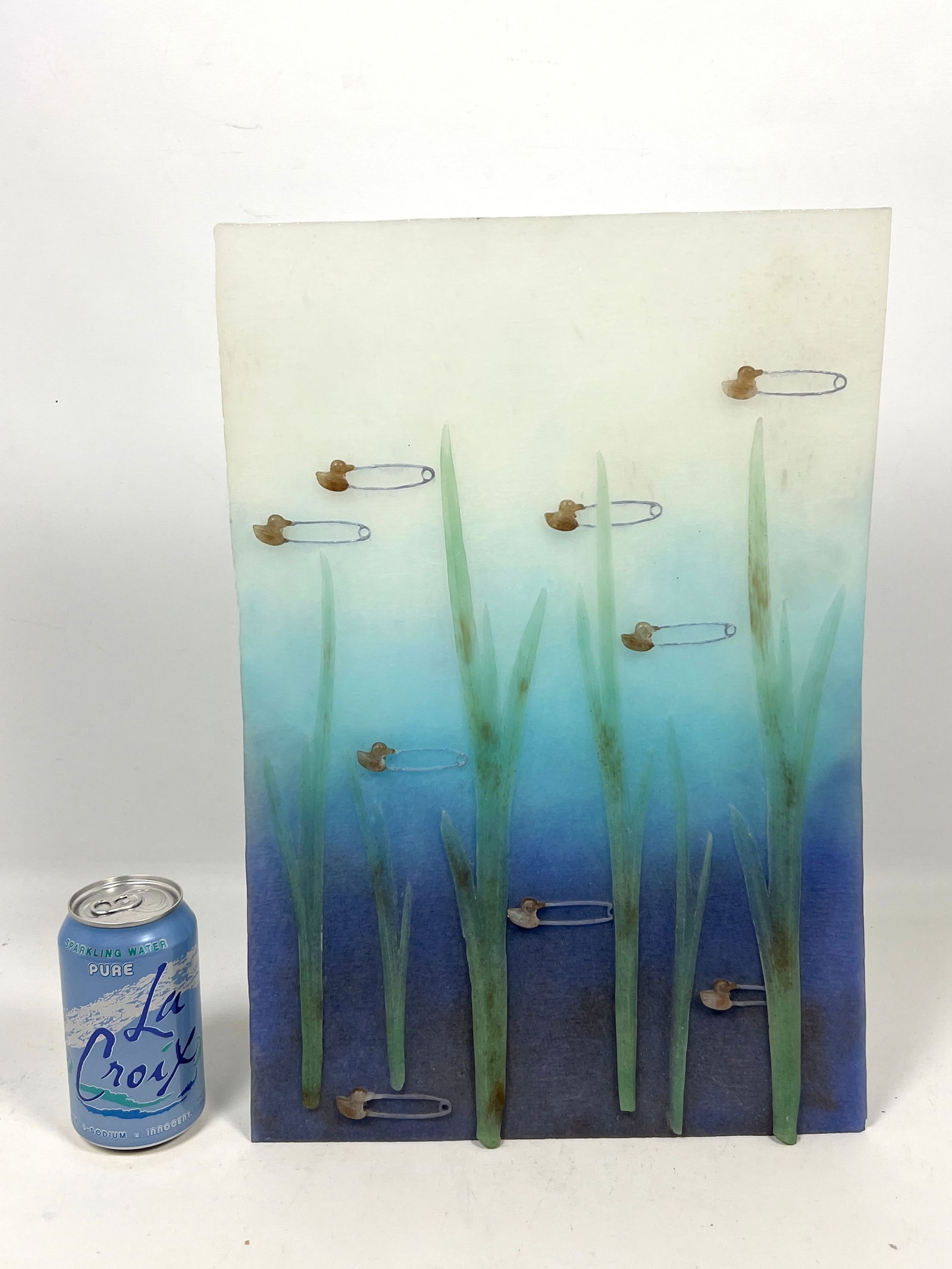 A vertical mixed-media artwork featuring tall green blades and scattered safety pins against a blue gradient background.