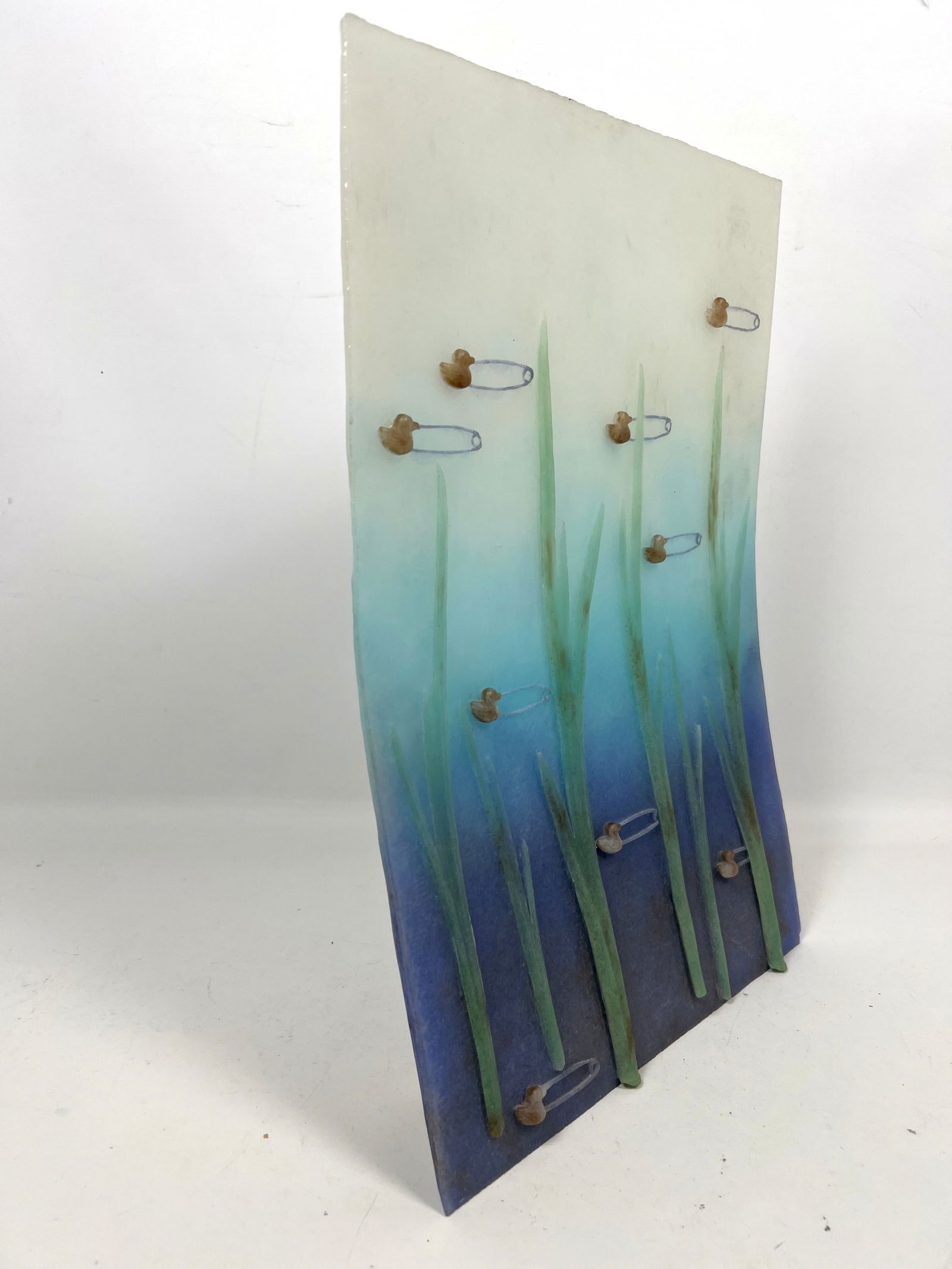 A vertical mixed-media artwork featuring tall green blades and scattered safety pins against a blue gradient background.